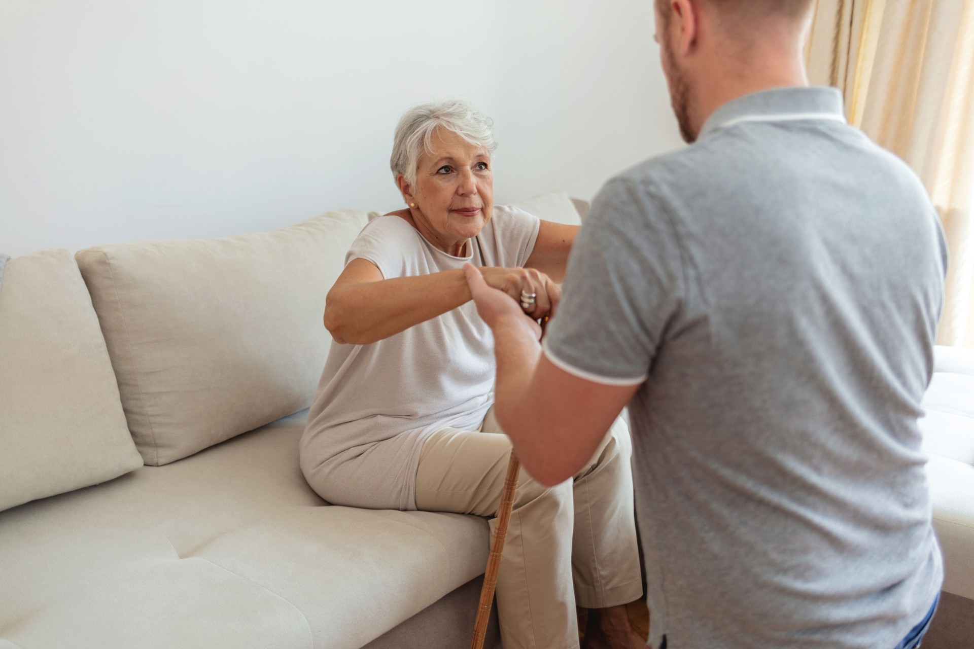Female home care supporting old woman to stand up from the sofa at car. Photo of male professional caregiver taking care of elderly woman at home during the day.